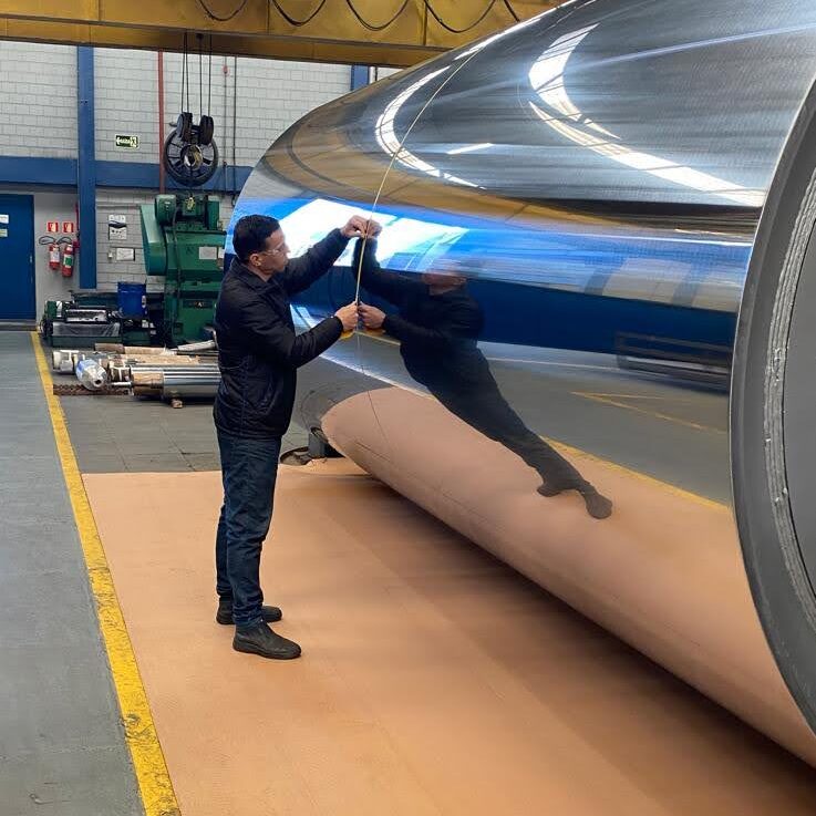 Homem realiza medição em cilindro metálico com superfície altamente polida dentro de uma indústria, ilustrando controle de qualidade para prevenção de corrosão eletroquímica.