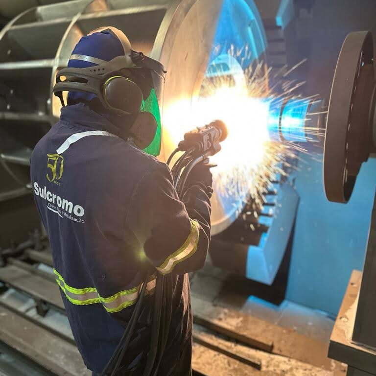 Técnico realiza processo de metalização com jato térmico em componente industrial para proteção contra corrosão eletroquímica, utilizando equipamentos de segurança e uniforme da Sulcromo.